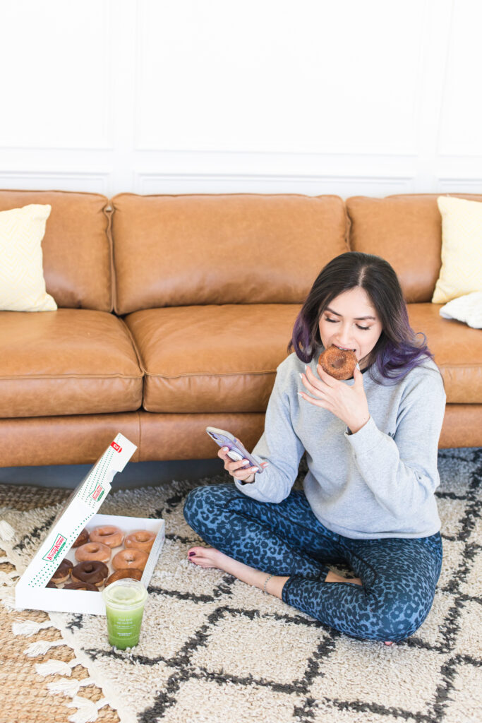 Woman eating donuts and a green juice cross legged on a rug on the floor next to the coach