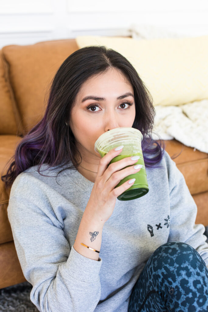 Woman sitting on the floor leaning against the coach drinking a green juice